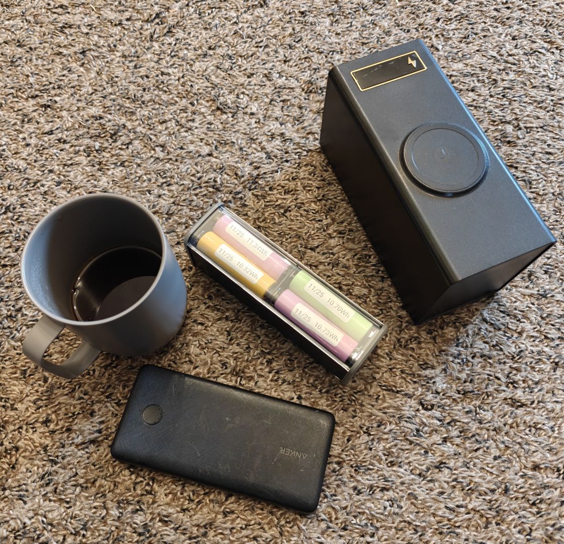 An Anker branded 10,000Mah (24Wh) battery bank sitting next to an aliexpress 86Wh battery bank with a clear back showing 8 reclaimed 18650s sitting next to a very large aliexpress over 400Wh battery bank containing 40 18650s. A mug of coffee is also shown.